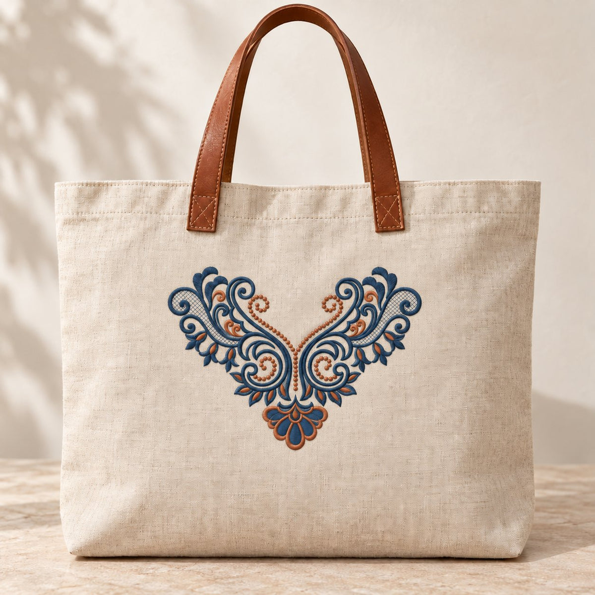Natural linen tote with leather handles and a smaller blue-and-copper V-shaped acanthus embroidery on the front.