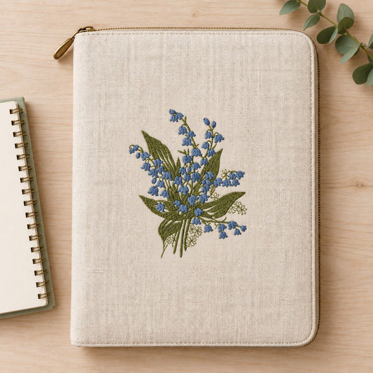 Closed natural linen zip folio beside a notebook, showing a blue bell-flower bouquet with sage foliage.
