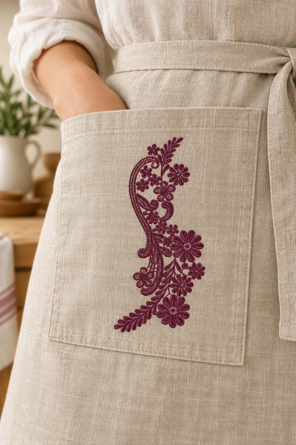 Photorealistic paisley wildflower spray machine embroidery in deep mulberry thread on a linen apron pocket, shown as one fabric-stitched motif