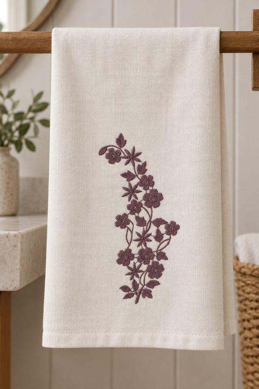 Linen guest towel with a single muted dark mulberry machine-embroidered petal and starflower vine motif on natural ivory linen.