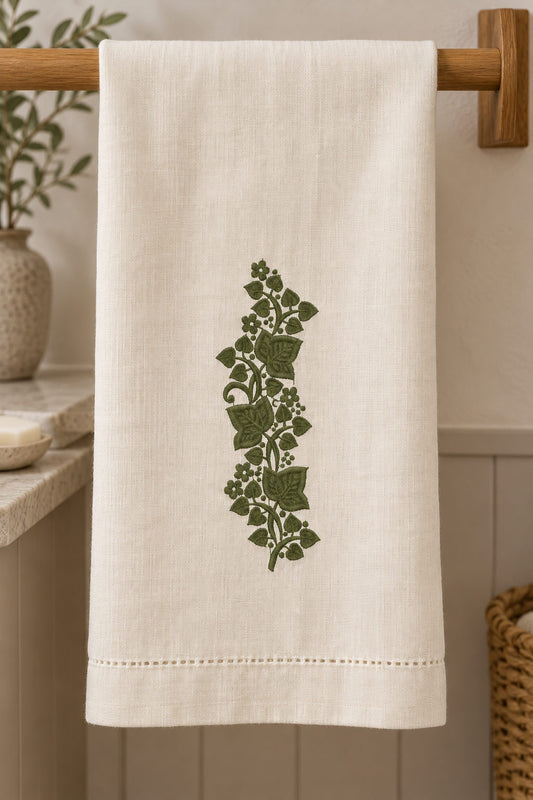 Linen guest towel with a single deep muted olive green machine-embroidered ivy leaf and blossom sprig motif on natural ivory linen.