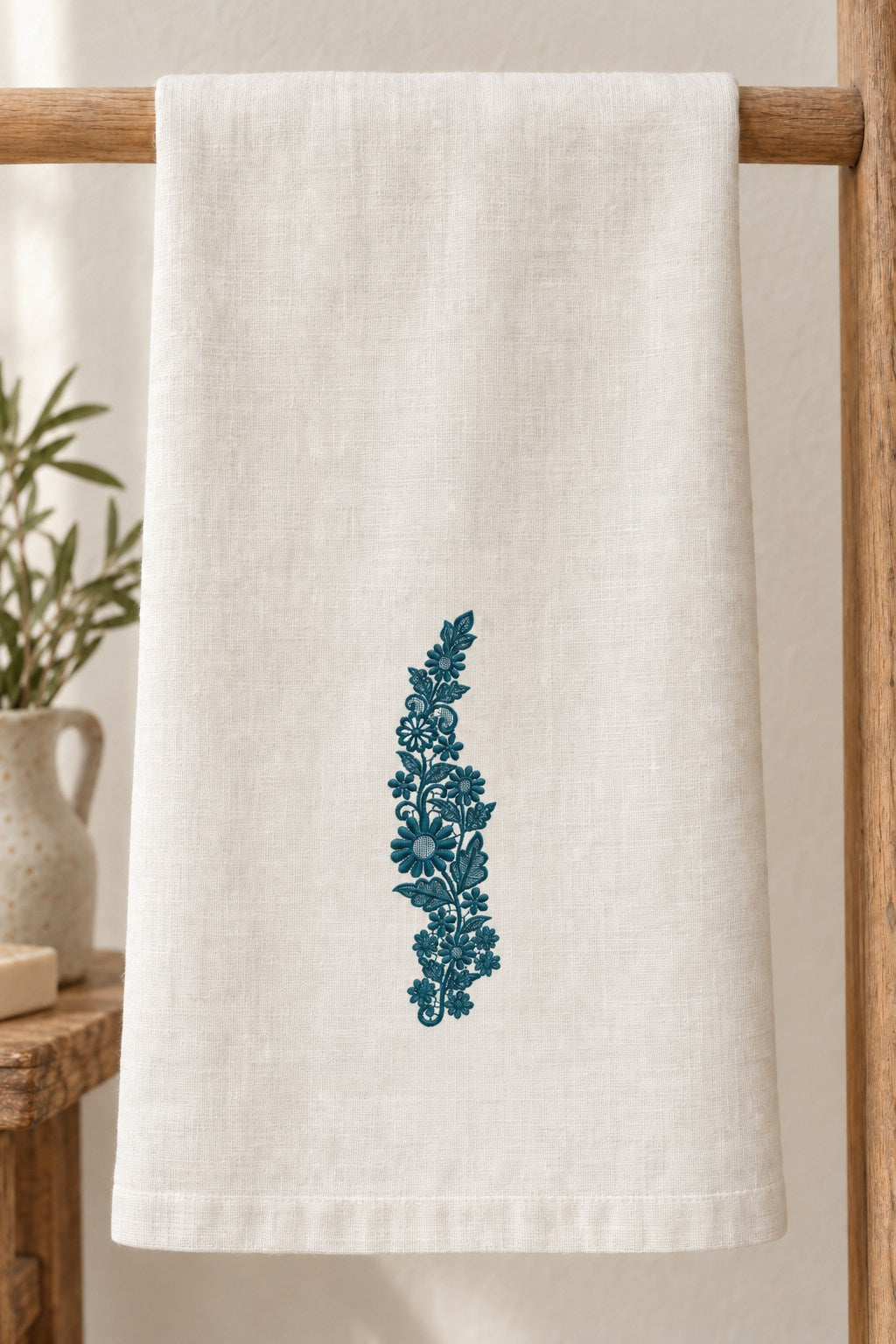 Styled linen hand towel mockup with a single deep muted petrol teal daisy meadow lace vine embroidered on pale linen; the complete motif has clear fabric around it.