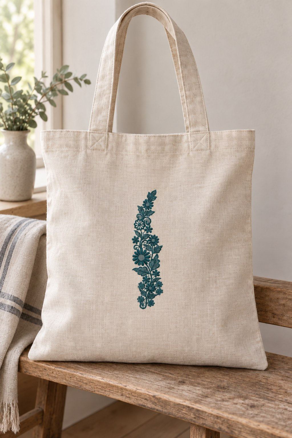 Styled linen tote bag mockup with a single deep muted petrol teal daisy meadow lace vine embroidered on pale linen; the complete motif has clear fabric around it.