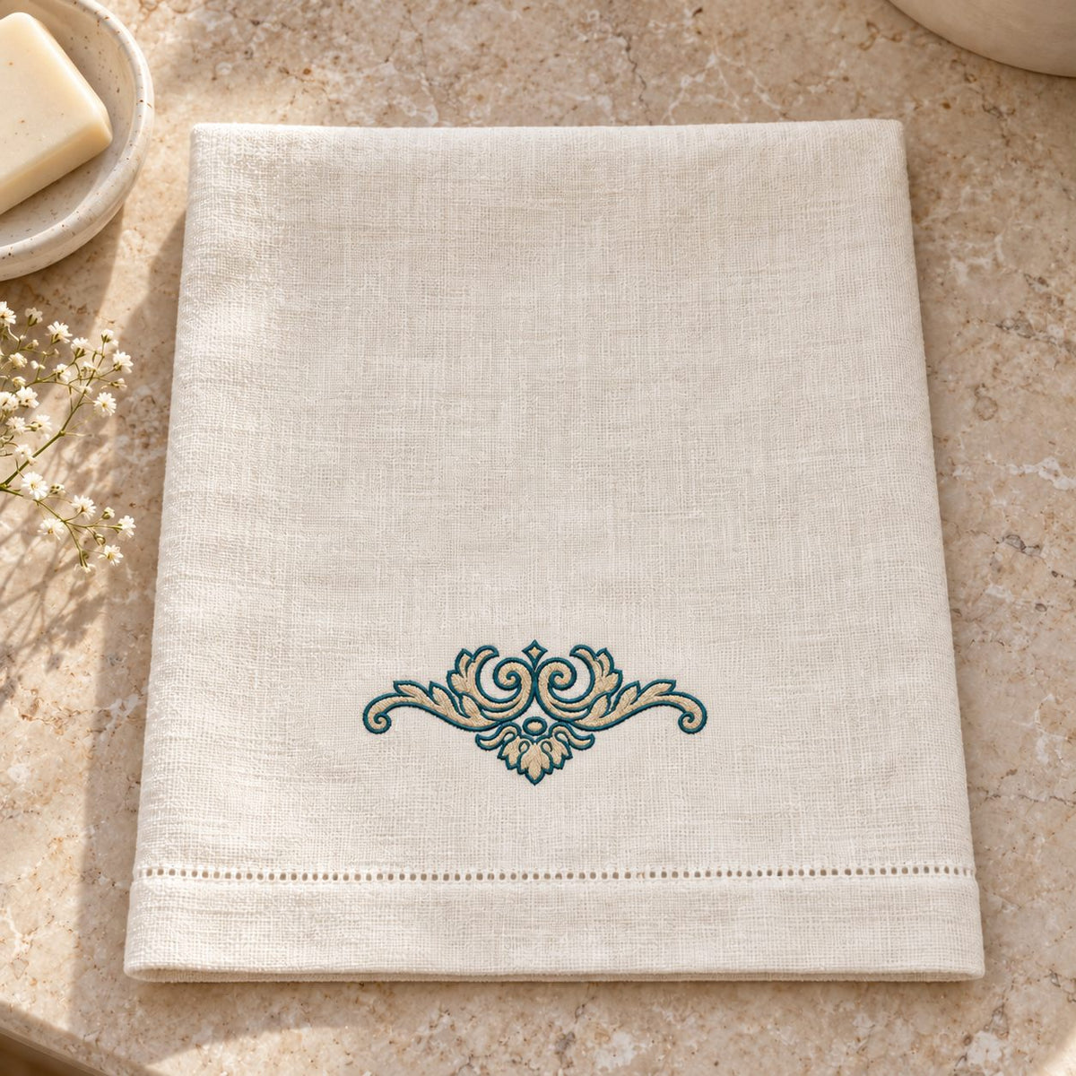 Oatmeal linen guest towel with a small parchment-filled scroll crest outlined in petrol above the lower hem.