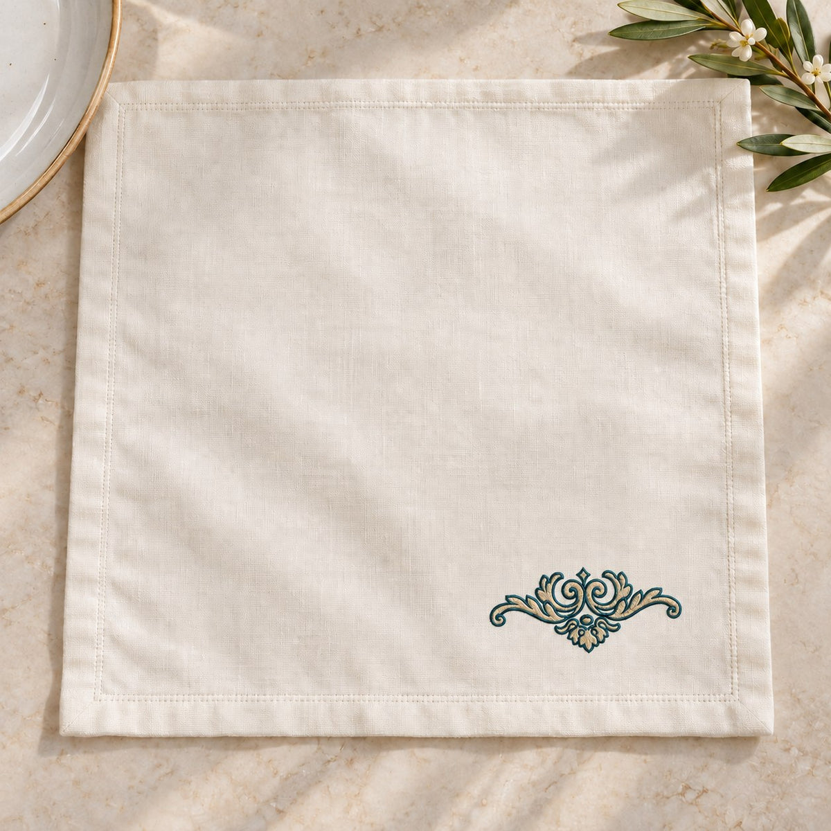 Ivory dinner napkin with a small two-color scroll crest near one corner, beside simple tableware.