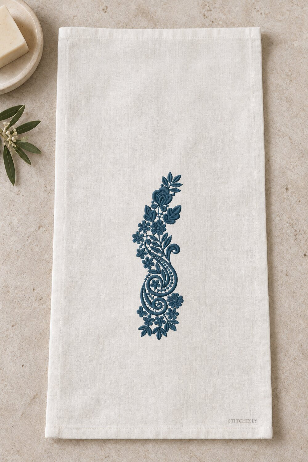 Generated usage mockup of a linen guest towel with the rose and twin paisley spray machine embroidery motif in one deep muted petrol blue thread color.