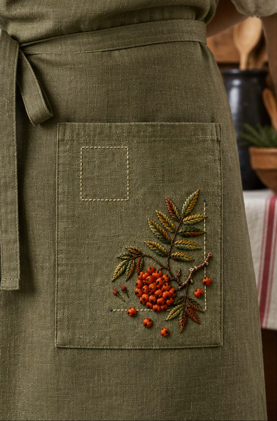 Green garment pocket mockup featuring the Rowan Pocket Corner hand embroidery design. Rowan berries and branching leaves occupy a lower-right corner beneath a clear label square.