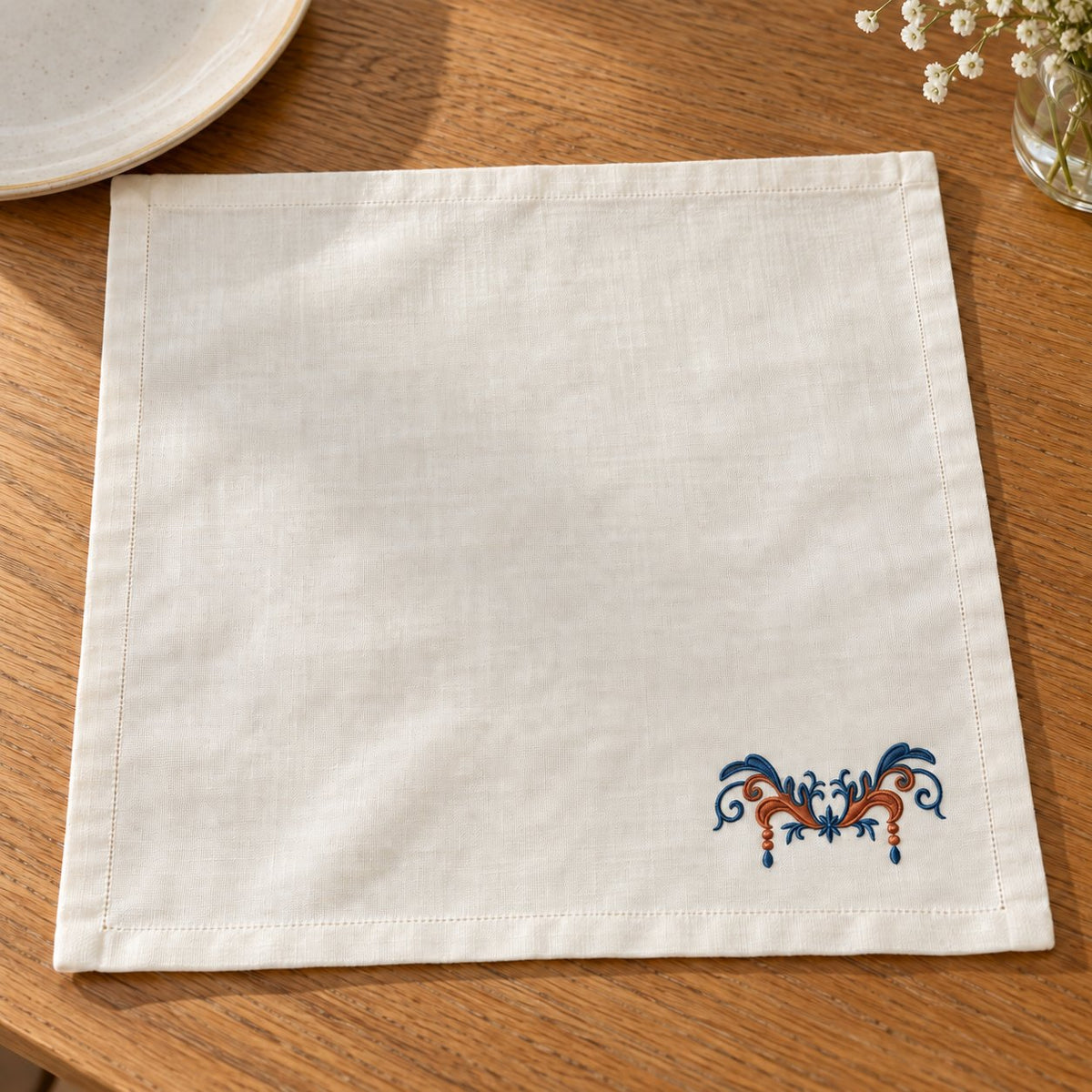 Linen napkin with a small terracotta-and-midnight acanthus swag near the corner on a wooden table.