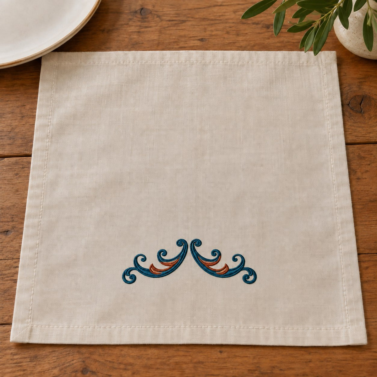 Twin Curl Acanthus Swag embroidered in petrol teal and muted copper on a linen table napkin, showing the complete two-color motif.